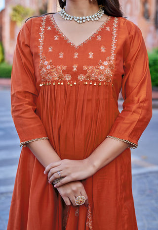 A woman wearing a rust-orange cotton kurta with gold embroidery on the neckline and sleeve cuffs, styled with a silver-toned statement necklace and rings.