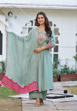 A woman models a mint green traditional Indian salwar kameez suit with a matching dupatta in an outdoor courtyard setting. The kurta features intricate floral embroidery on the chest and pink patterned cuffs, while the dupatta has pink border detailing.