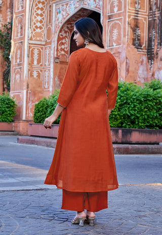 A woman wearing a rust-orange kurta and matching pants, photographed from behind in front of a historic terracotta-colored archway with green hedges.