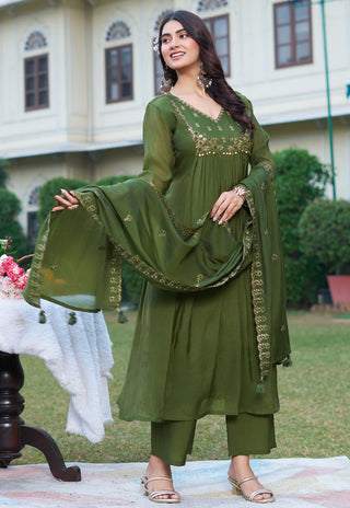 A woman models an olive green traditional Indian salwar kameez suit with gold embroidery, standing outdoors in front of a building with green lawns.