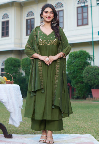 A woman stands outdoors wearing an olive green traditional outfit with gold embroidery and a matching draped scarf. The background shows a grassy lawn, green shrubs, and a cream-colored building.