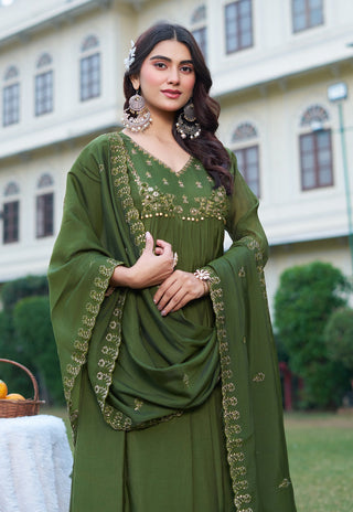 A woman wearing an olive green traditional outfit with gold embroidery and a matching dupatta, standing outdoors in front of a large cream-colored building.